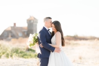 Groom kissing bride who has eyes closed while holding a wedding bouquet on a beach in Sandy Hook, NJ