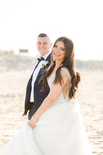 Bride with Full Hair & Makeup On Sandy Hook NJ beach smiling at camera holding groom's hand